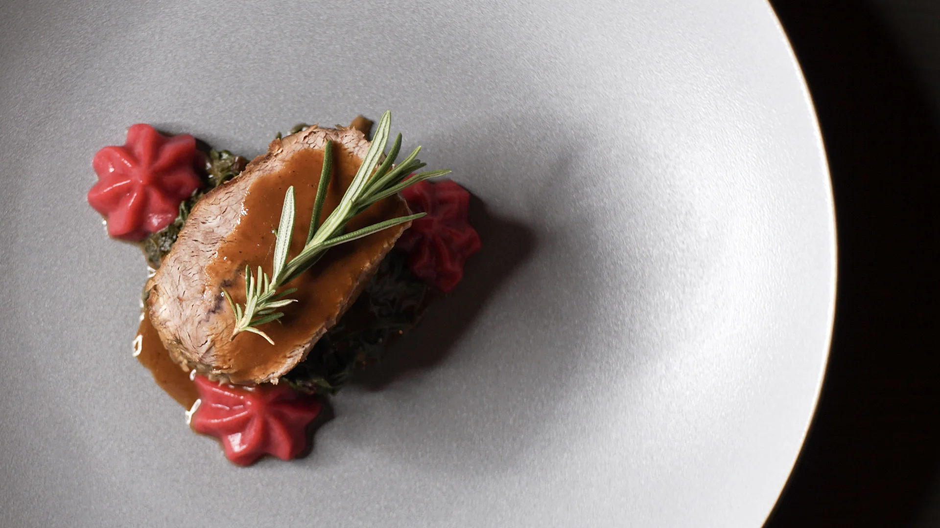 Gourmetgericht mit Fleisch, Sauce, Kräutern und rotem Gemüse auf grauem Teller