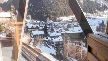 Blick aus einem Holzsauna-Fenster auf schneebedecktes Dorf und Berge