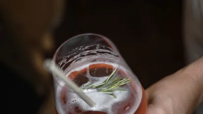 Hand hält Glas mit rotem Cocktail und Rosmarinzweig