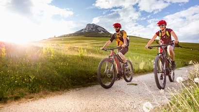 Zwei Radfahrer mit Helmen fahren auf einem Weg durch grüne Hügel bei Sonnenschein