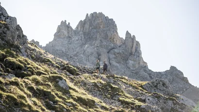 Zwei Wanderer auf felsigem Bergpfad mit spitzen Gipfeln im Hintergrund